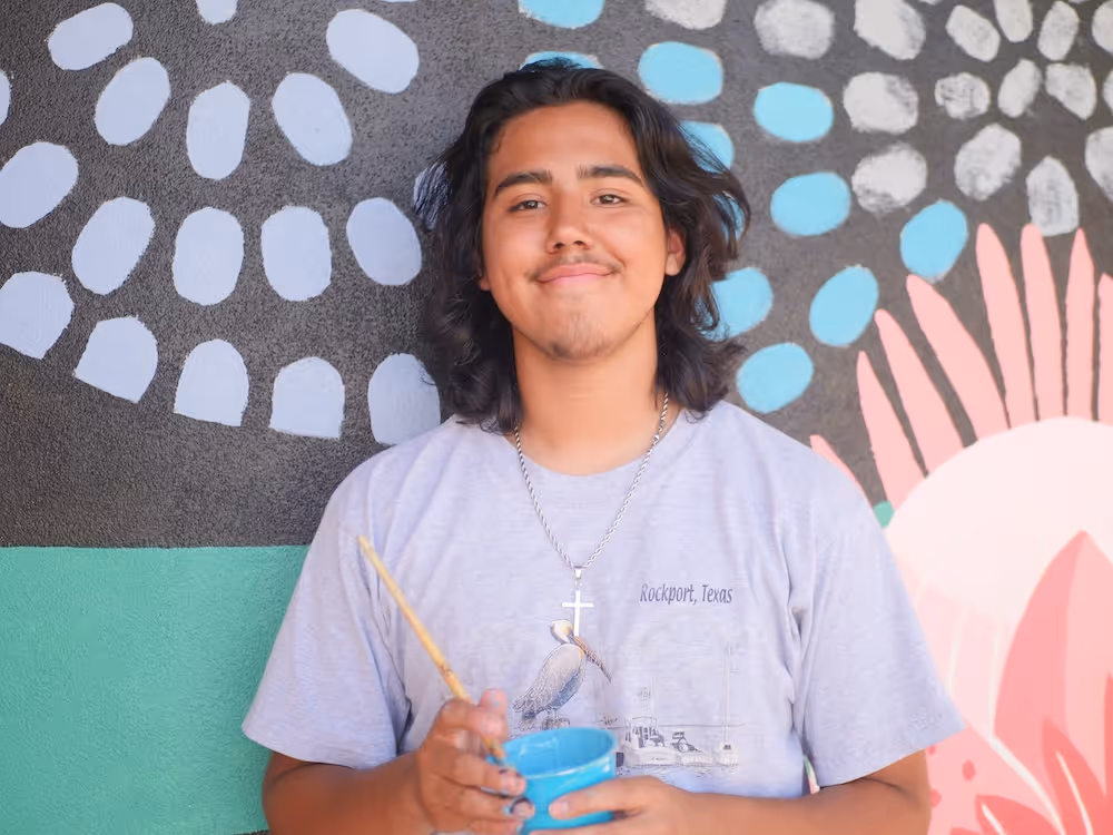 Smiling young man holding a paintbrush and blue paint cup in front of a colorful mural.