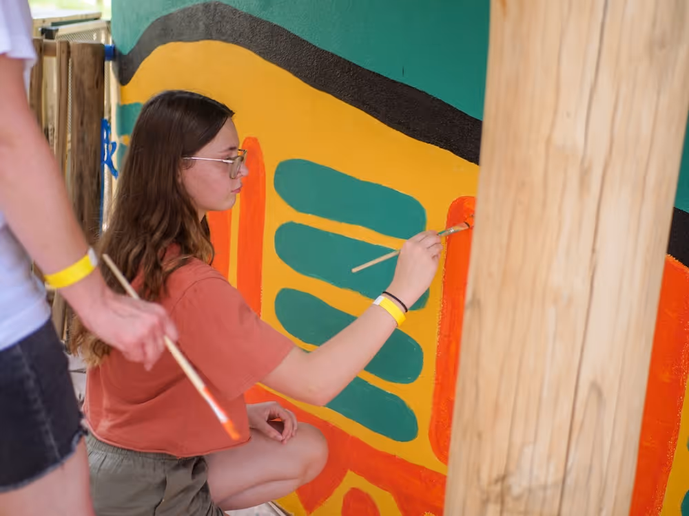 Young woman with glasses painting colorful abstract shapes on a wall while kneeling, with another person holding a paintbrush nearby.