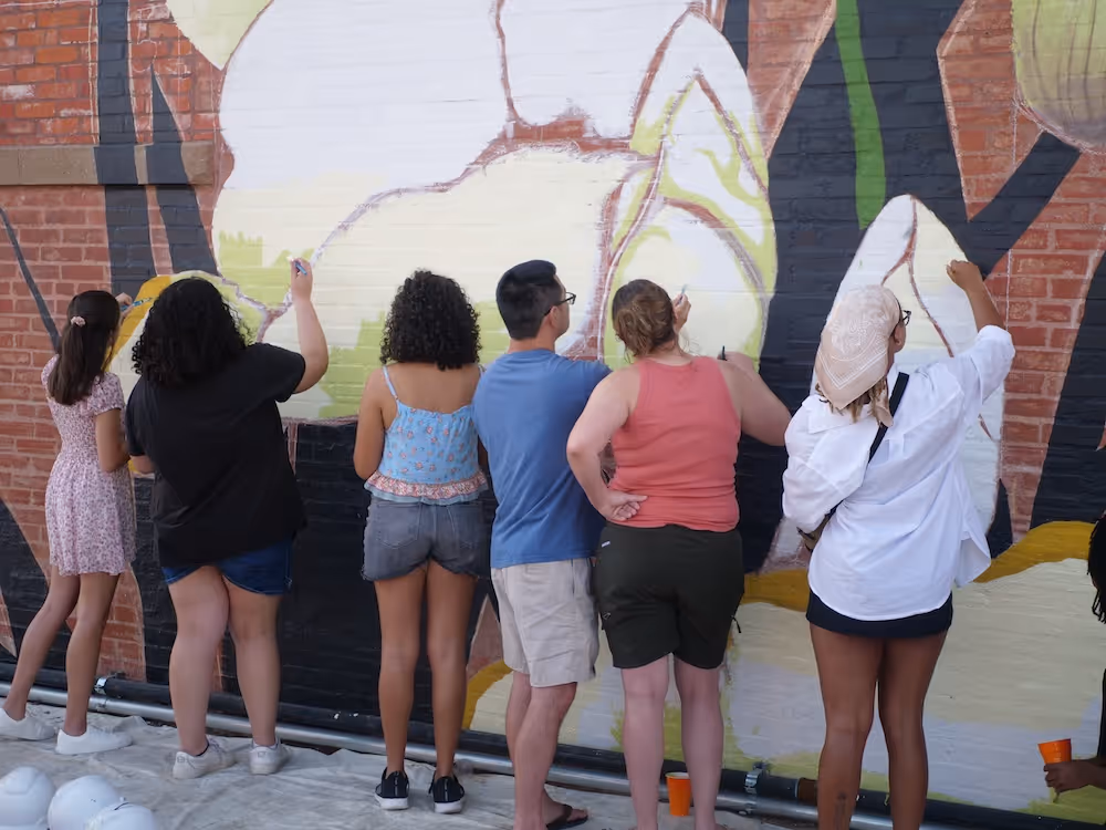 Six people painting a large mural on a brick wall outdoors, standing on a covered ground.