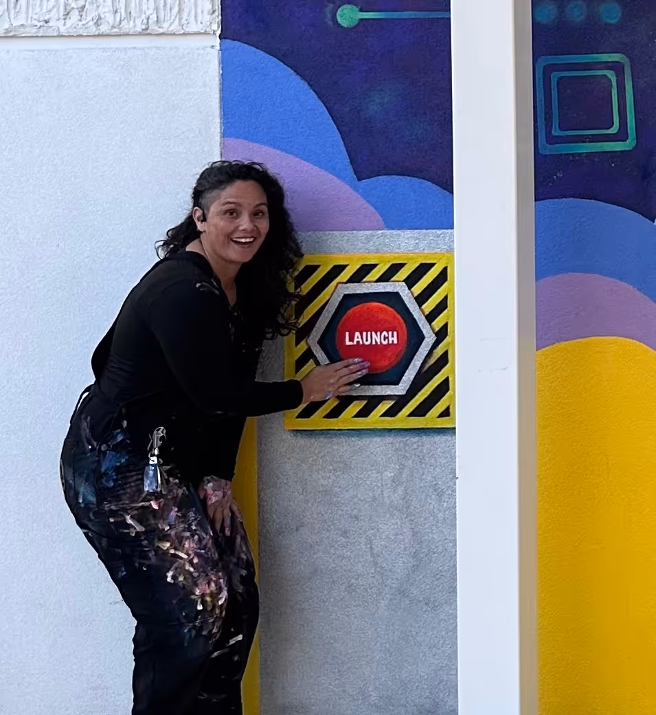 Woman with curly hair smiling and pressing a large red button labeled 'LAUNCH' on a colorful wall.
