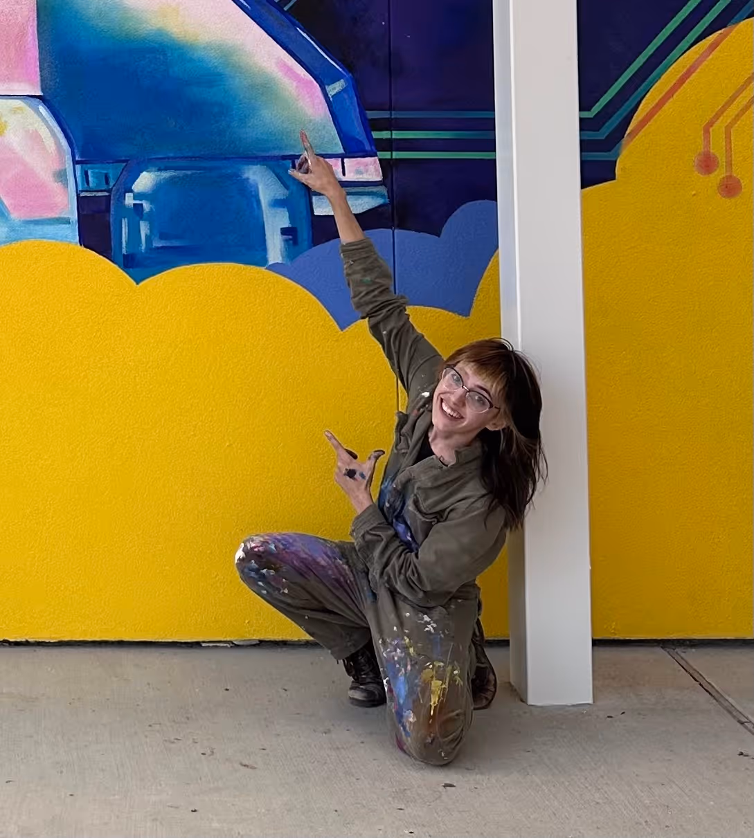 Smiling woman with glasses in paint-splattered coveralls poses kneeling and pointing at a colorful mural on a yellow wall.