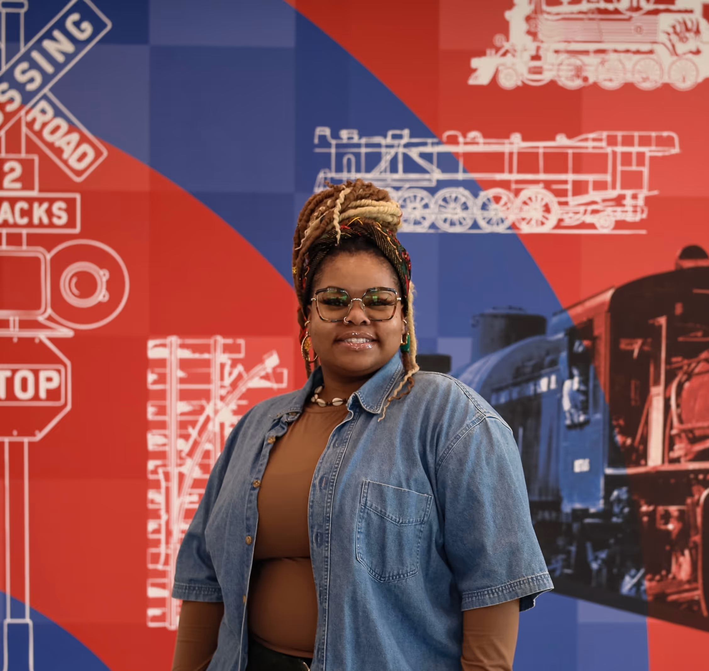 Woman with glasses and braided hair in a denim shirt stands in front of a colorful wall with train and railroad crossing illustrations.