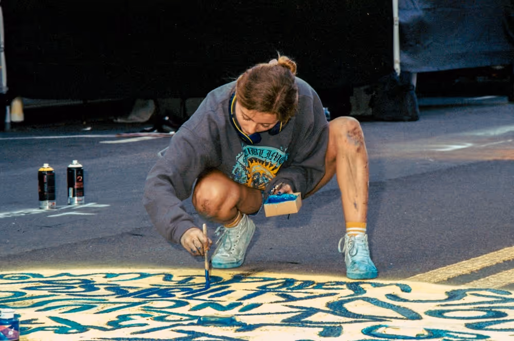 Young woman crouched on the ground painting blue calligraphy on a street surface, with paint cans nearby.