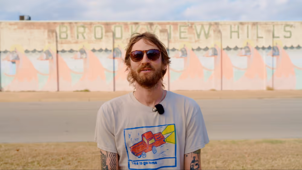 Bearded man wearing sunglasses and a white t-shirt with a red truck graphic standing outdoors in front of a wall with faded 'Brookview Hills' text.