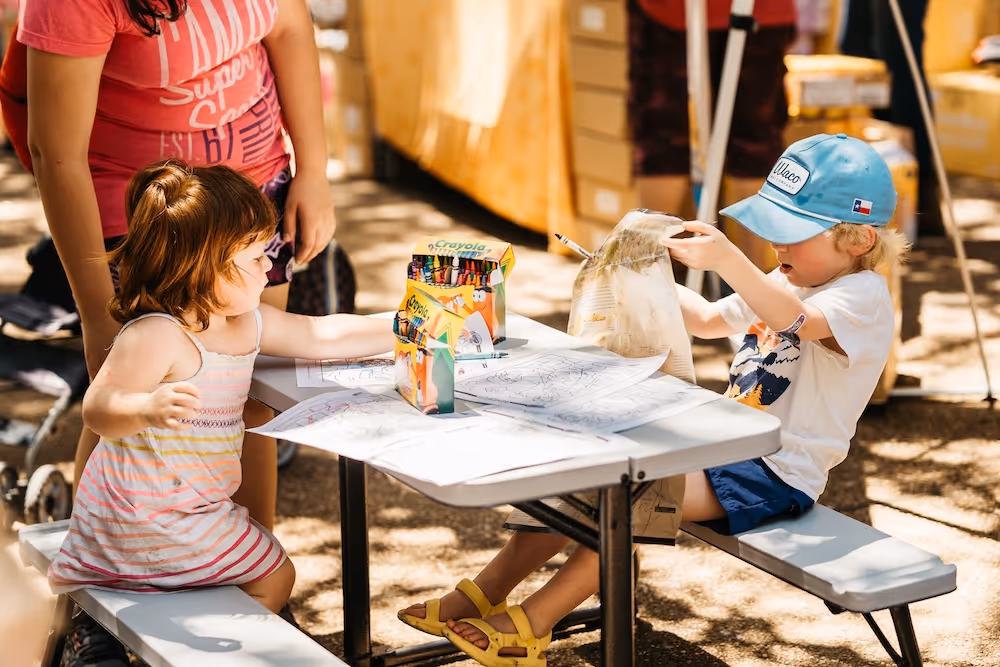 Two children at a picnic table coloring with crayons, a woman stands nearby outside in sunlight.
