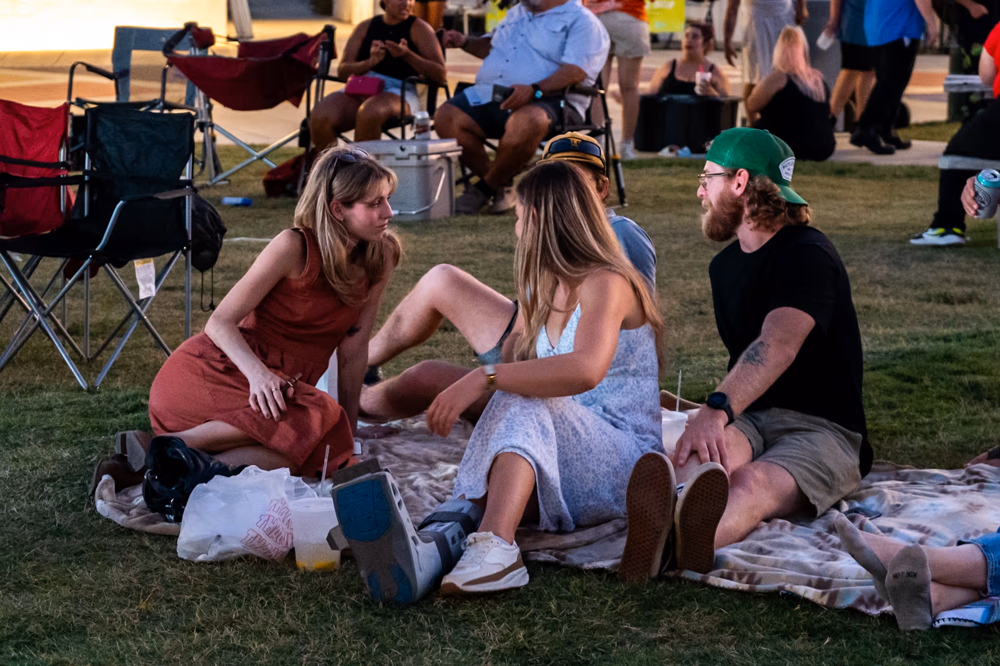 Group of four people sitting on a blanket on grass at an outdoor event, with one person wearing a medical boot.