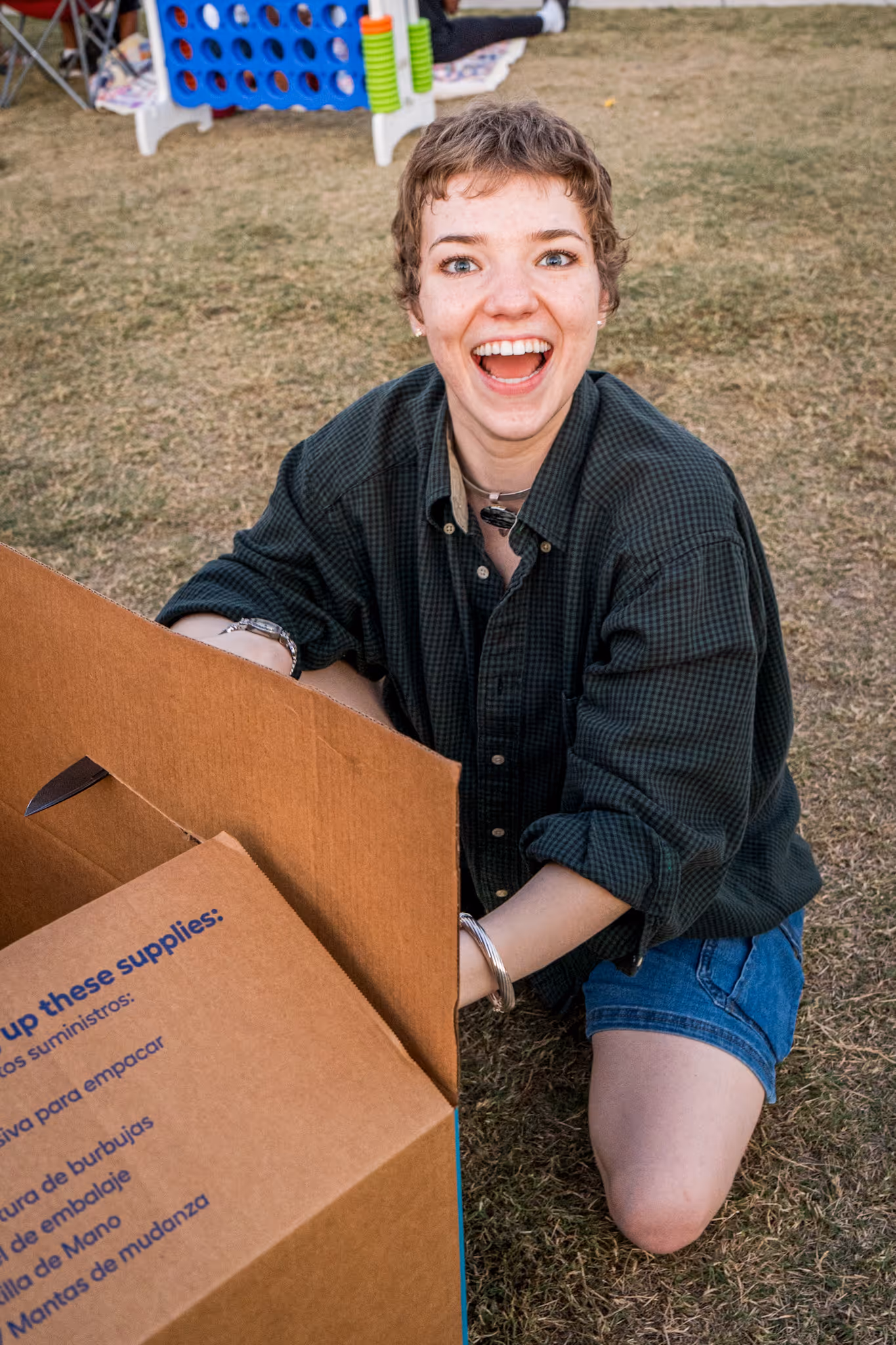 Smiling young person kneeling on grass, unpacking a large cardboard box outdoors.