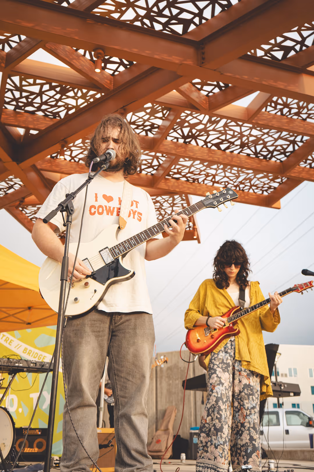 Two musicians performing outdoors under a decorative metal canopy, one singing and playing white electric guitar, the other playing red electric guitar.