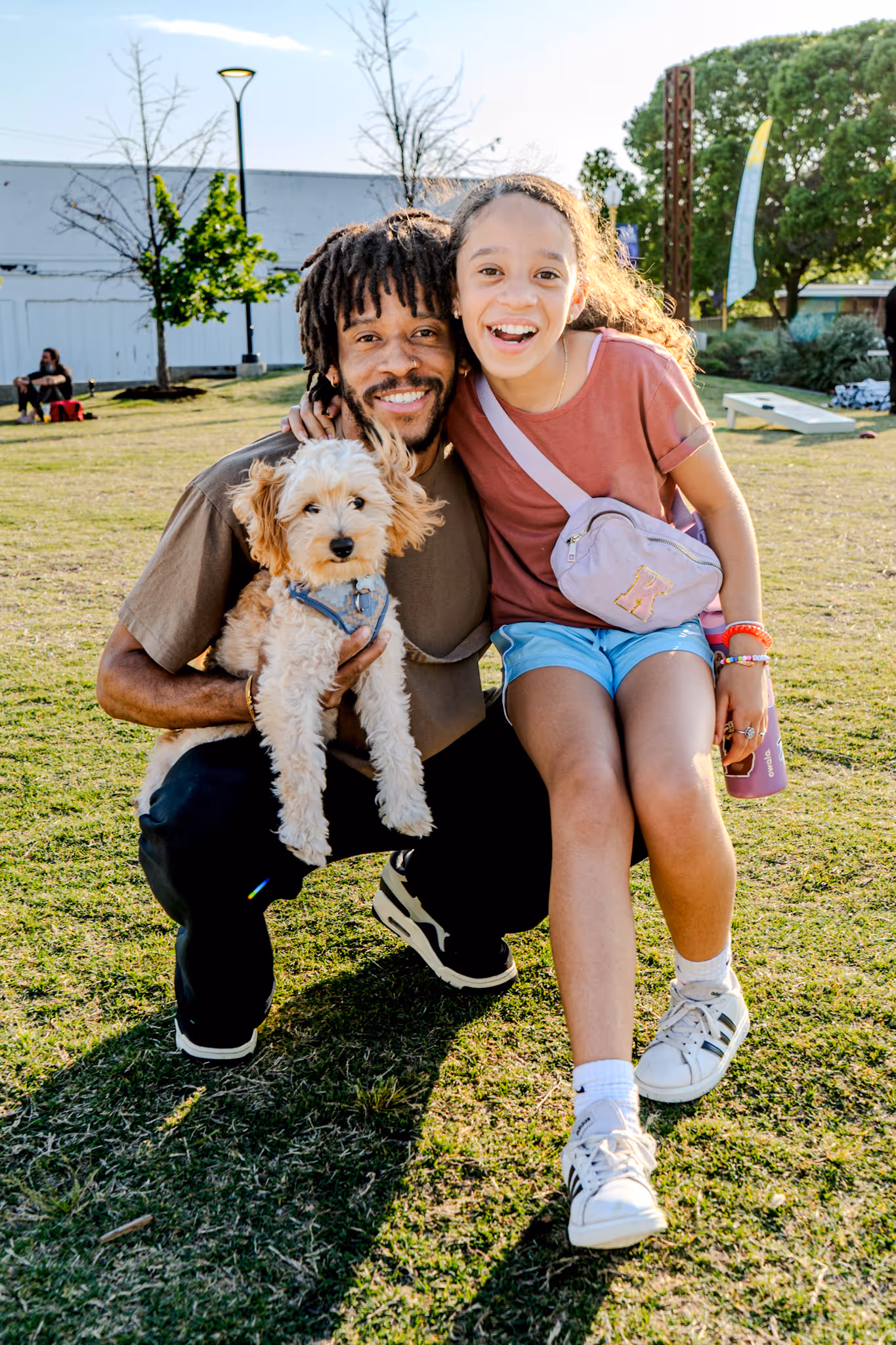 Smiling man crouching and holding a small fluffy dog next to a smiling girl sitting on grass in a park.