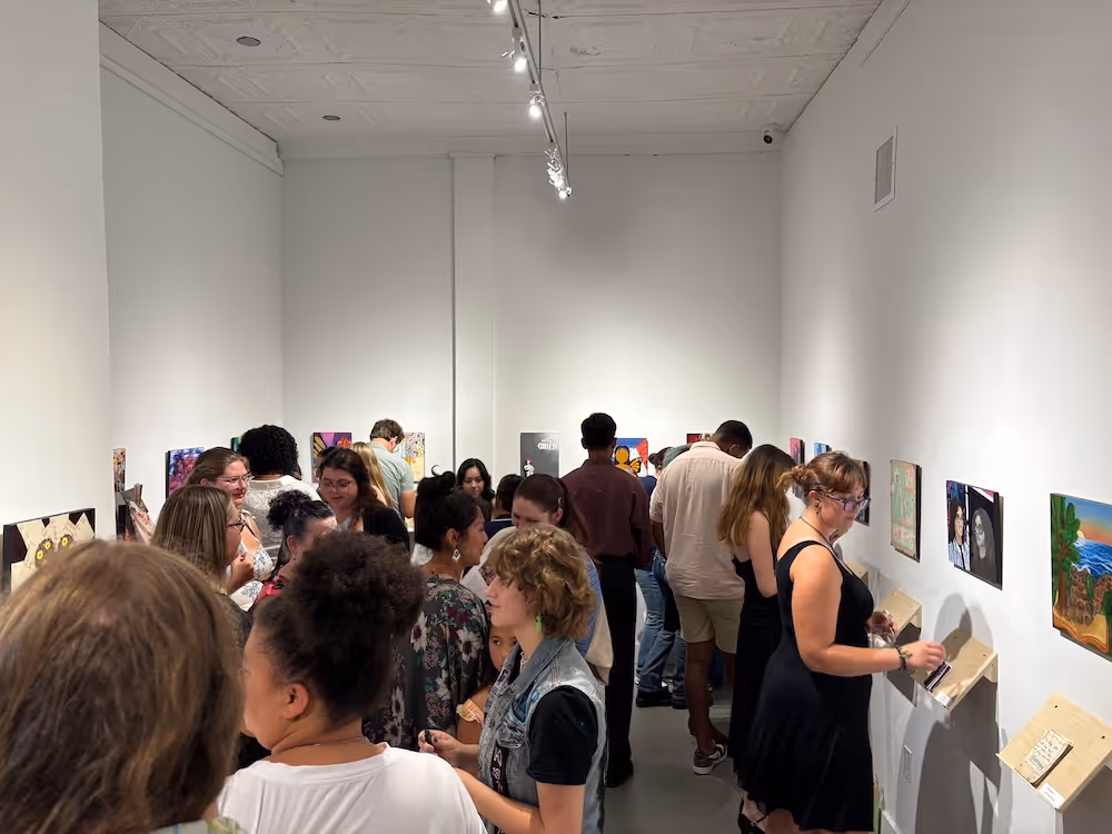 Crowd of people viewing and discussing artwork displayed on white gallery walls.