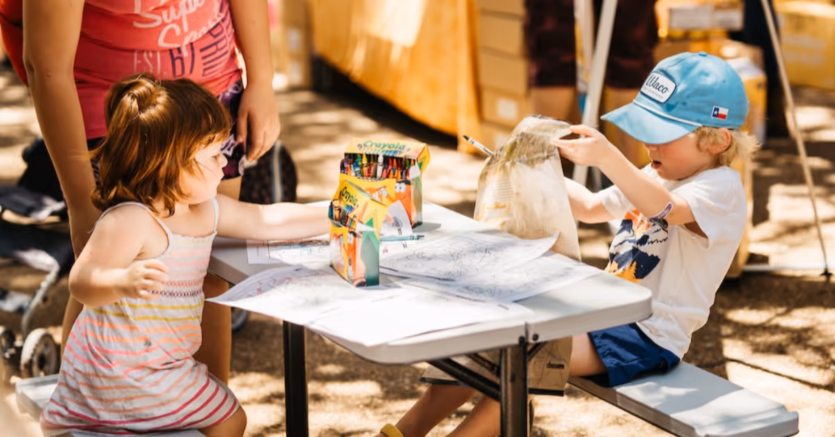 Two children sitting at a table coloring with crayons outdoors on a sunny day.