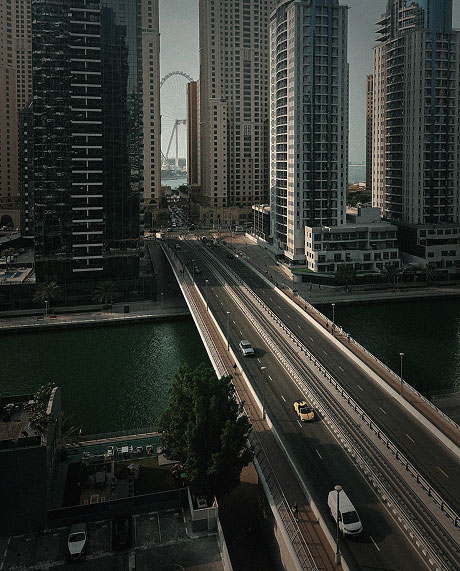 Bridge over water with cars, flanked by tall buildings, with a large Ferris wheel in the distance.