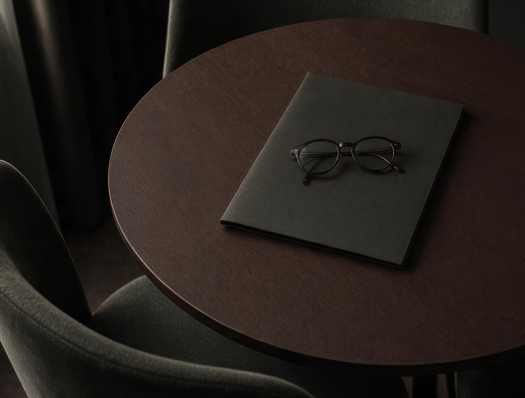 Round dark wood table with black folder and eyeglasses on top, surrounded by dark chairs.