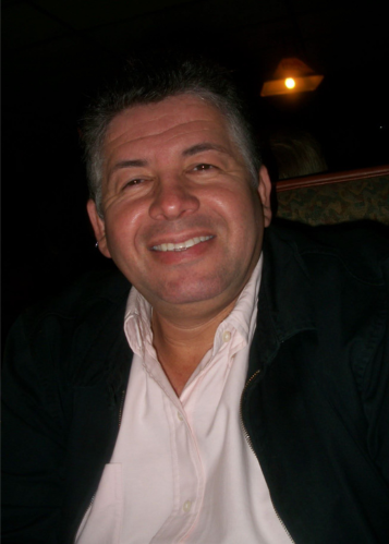 Smiling middle-aged man wearing a light pink shirt and dark jacket sitting in a dimly lit room.