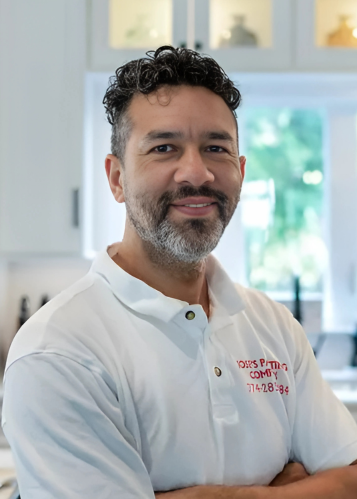 Man with curly hair and beard wearing a white polo shirt standing with arms crossed in a kitchen.