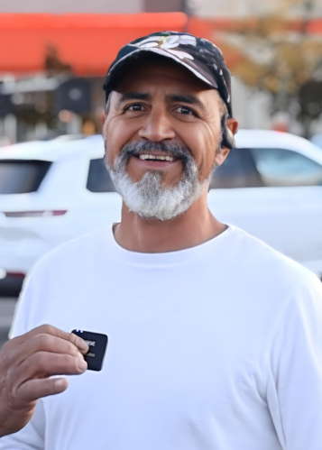 Smiling man with gray beard wearing a black cap and white shirt holding a car key fob outdoors.