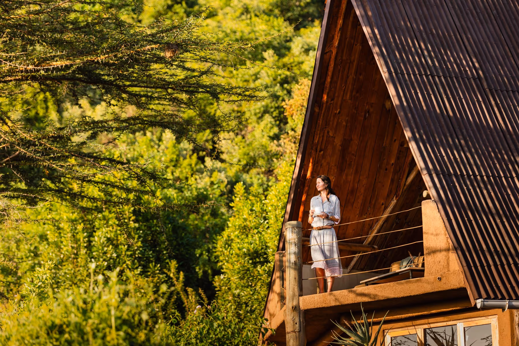 A peaceful moment of reflection as a guest enjoys a morning beverage on a private balcony overlooking lush green forest. The rustic architecture of the villa blends naturally with the surrounding landscape.