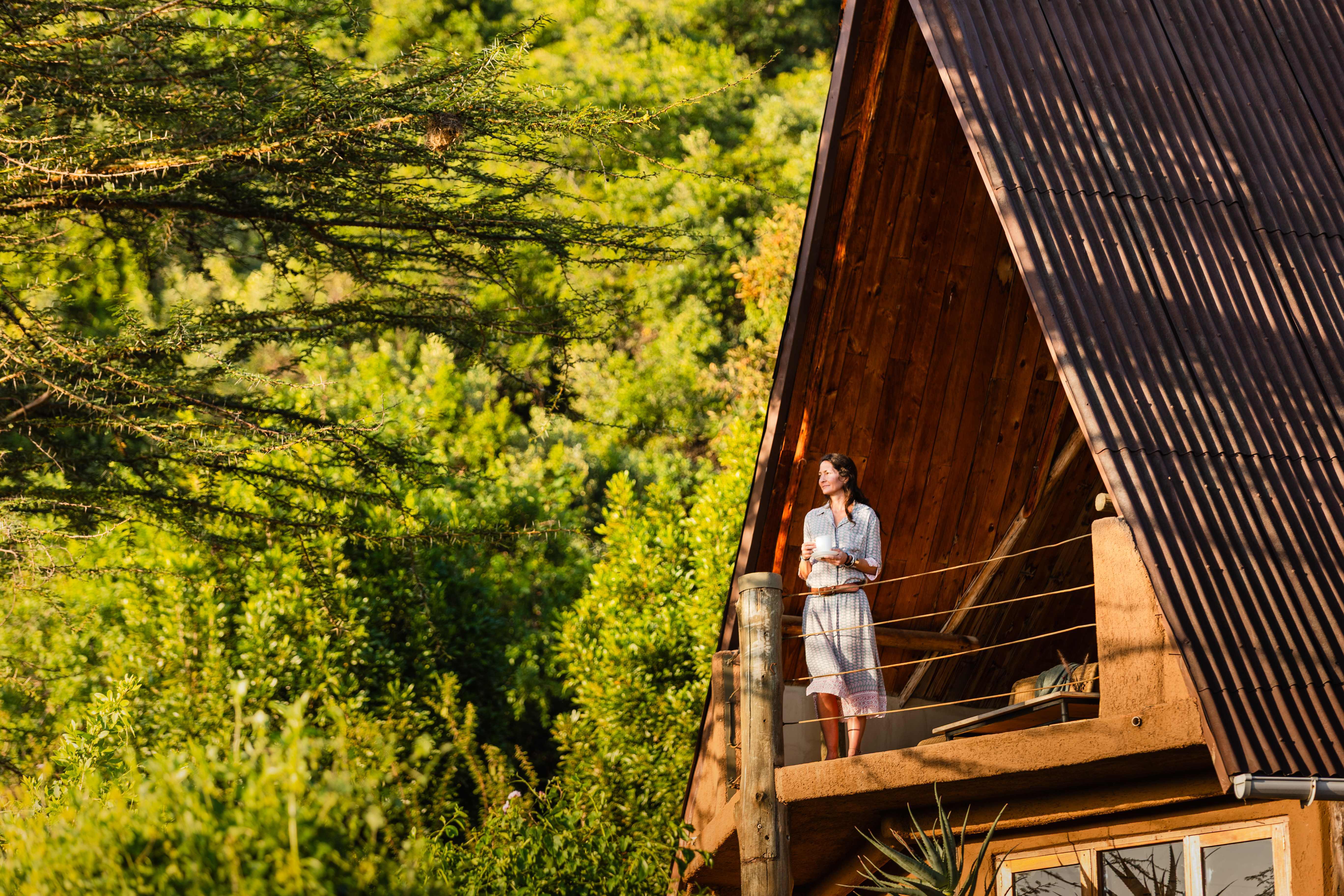 A peaceful moment of reflection as a guest enjoys a morning beverage on a private balcony overlooking lush green forest. The rustic architecture of the villa blends naturally with the surrounding landscape.