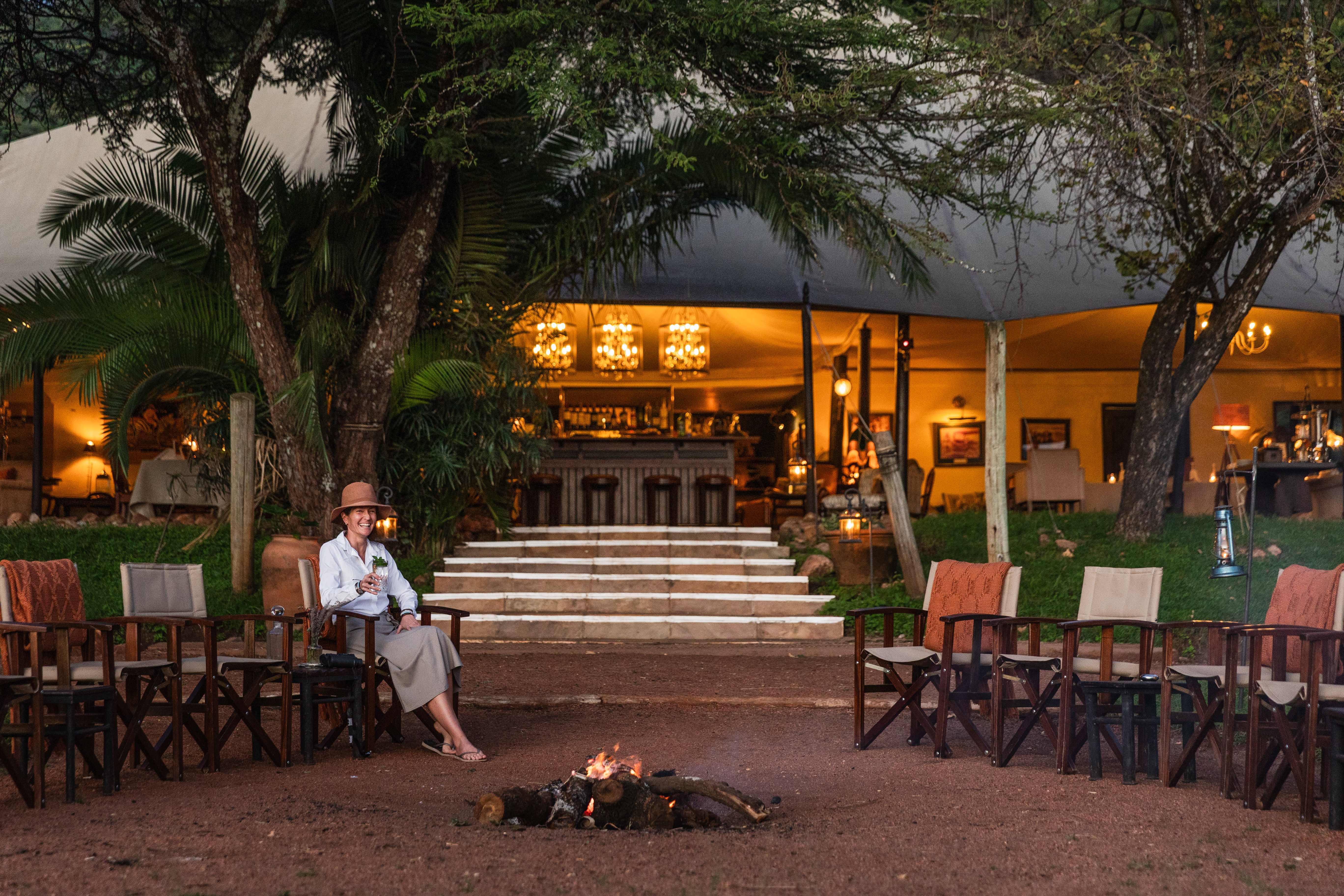A tranquil scene with a guest relaxing by a fire pit, surrounded by safari chairs in front of the warmly lit main tent. The ambiance reflects timeless elegance — lanterns, polished wood, and open canvas — evoking the golden era of safaris.