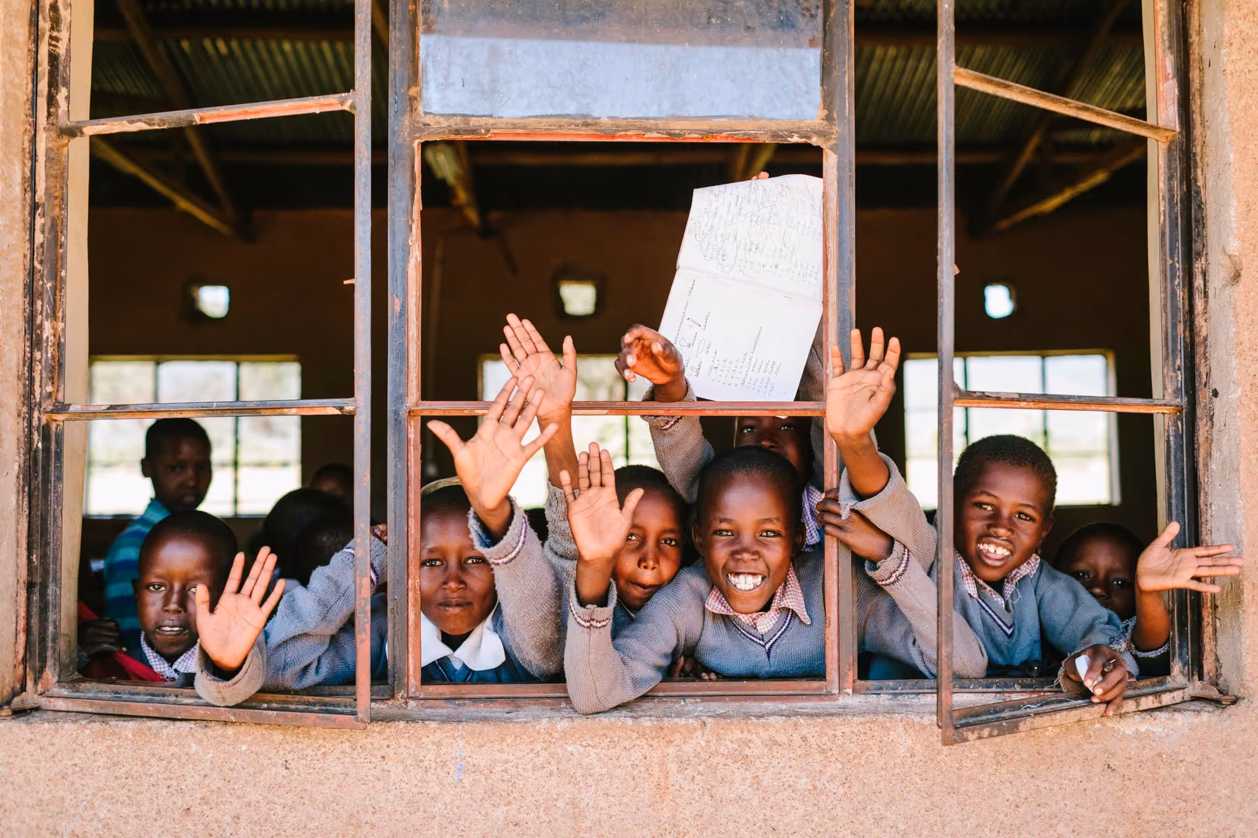 Maasai children smiling and waving from a classroom window during a community school visit supported by Cottar’s Safaris.
