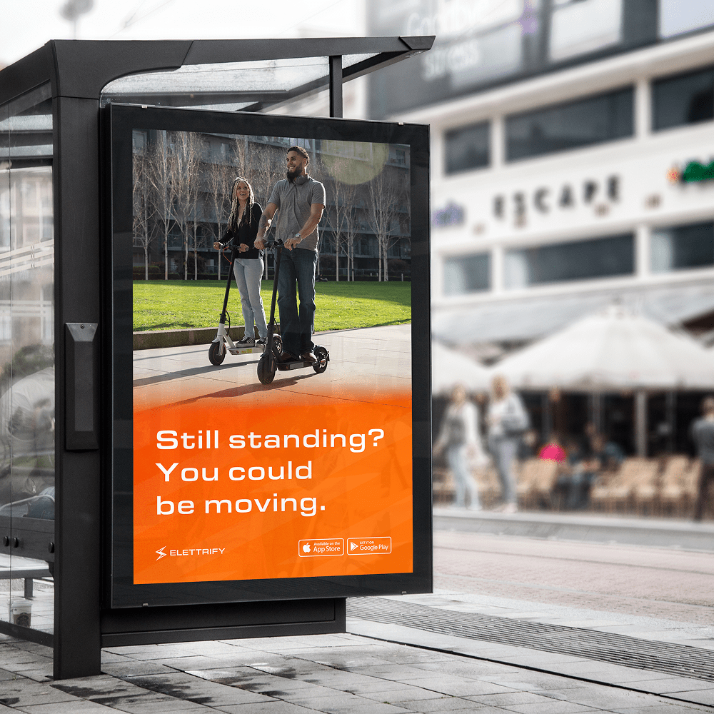 Elettrify e-scooter rental service bus stop advertisement featuring couple on electric scooters with 'Still standing? You could be moving.' campaign messaging