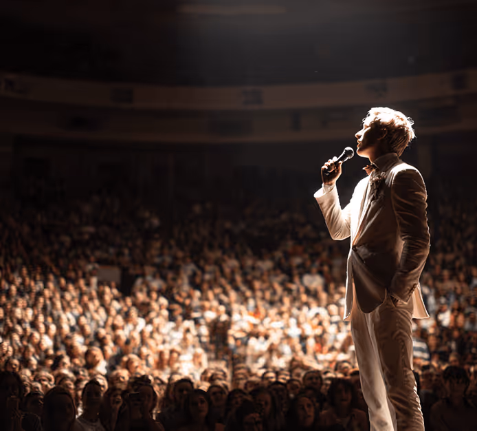 Performer singing on stage with large crowd in dimly lit concert venue