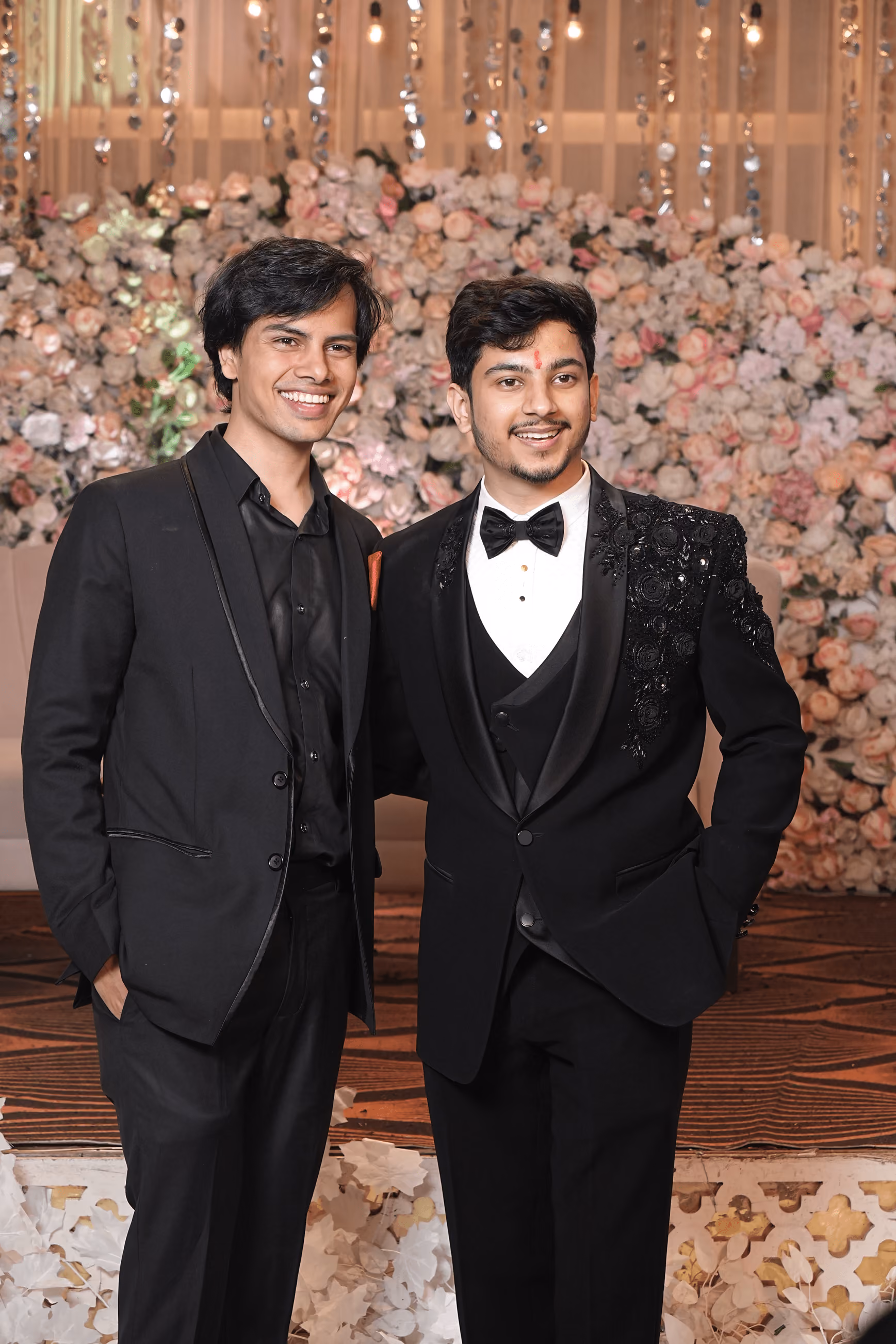 Two men in black suits smiling and posing in front of a floral backdrop with hanging lights.