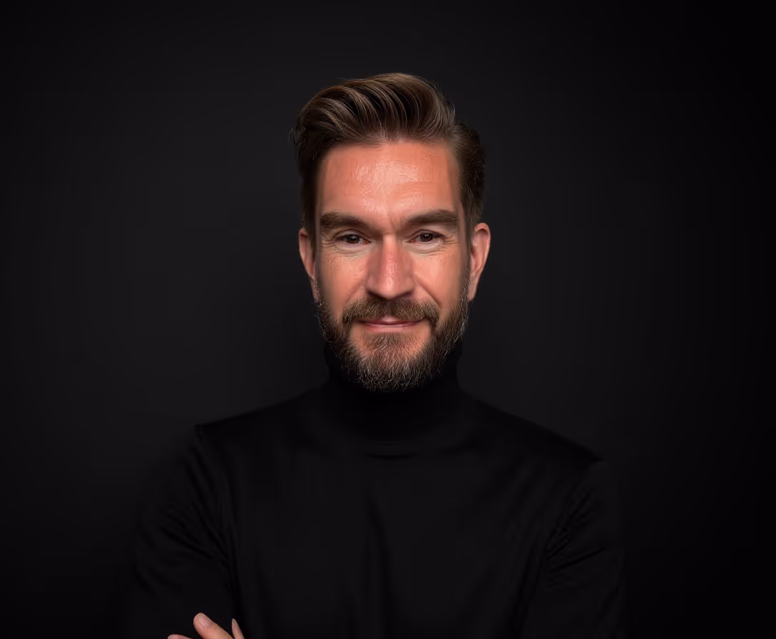 Porträt des Leadership Rebell, ein lächelnder Mann mit Bart und kurzem Haar in einem schwarzen Rollkragenpullover vor einem schwarzen Hintergrund.