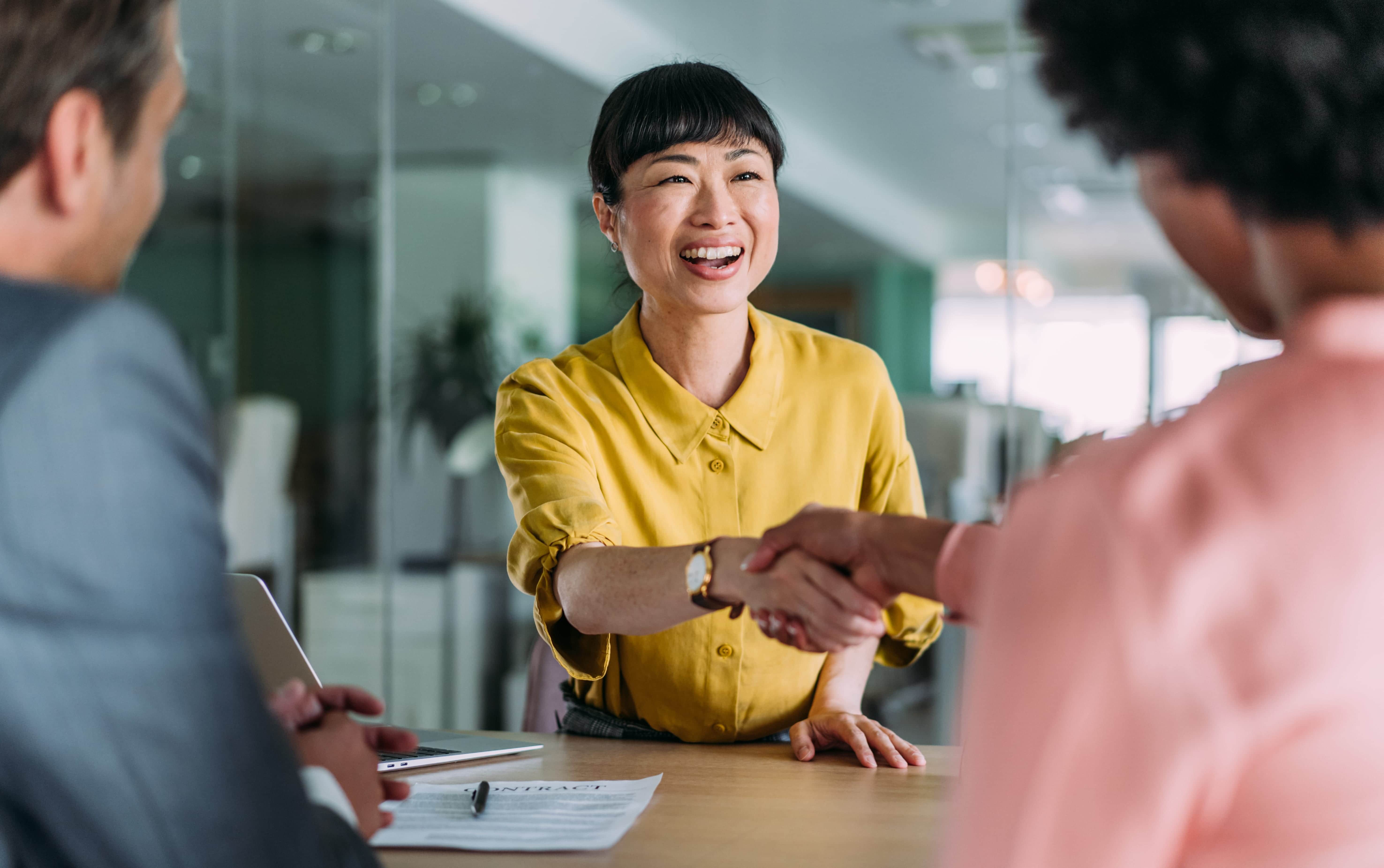 Smiling woman in yellow shirt shaking hands with another person across a table in an office setting.