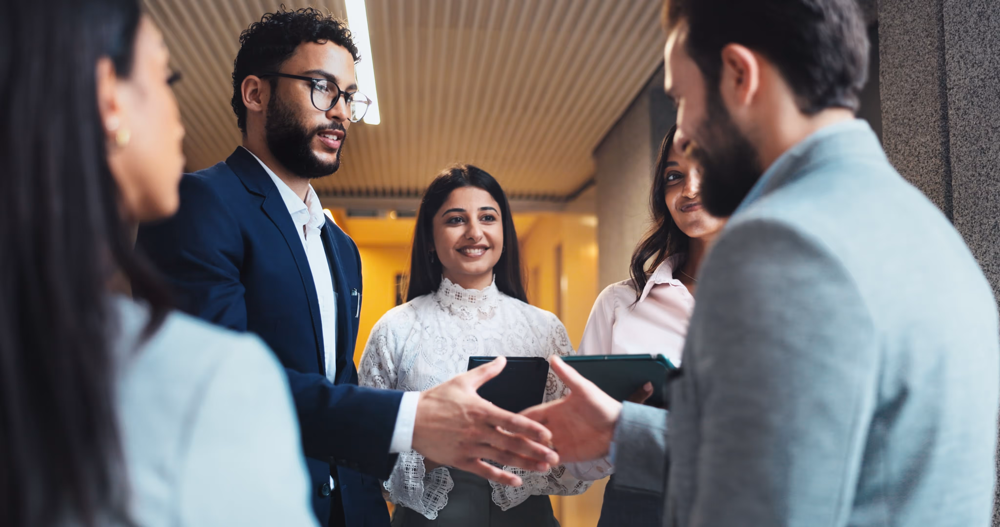 Two men shaking hands in a business setting while three women look on and smile.