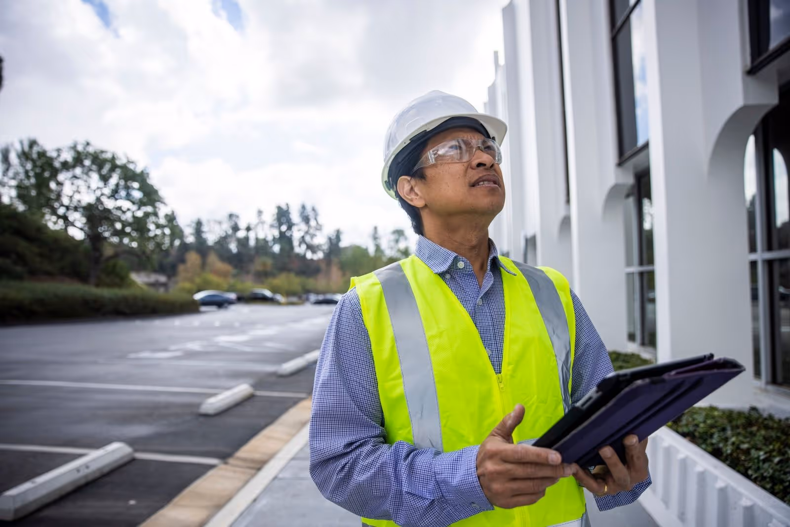 Construction worker in safety vest and hard hat holding a tablet outside a building site.