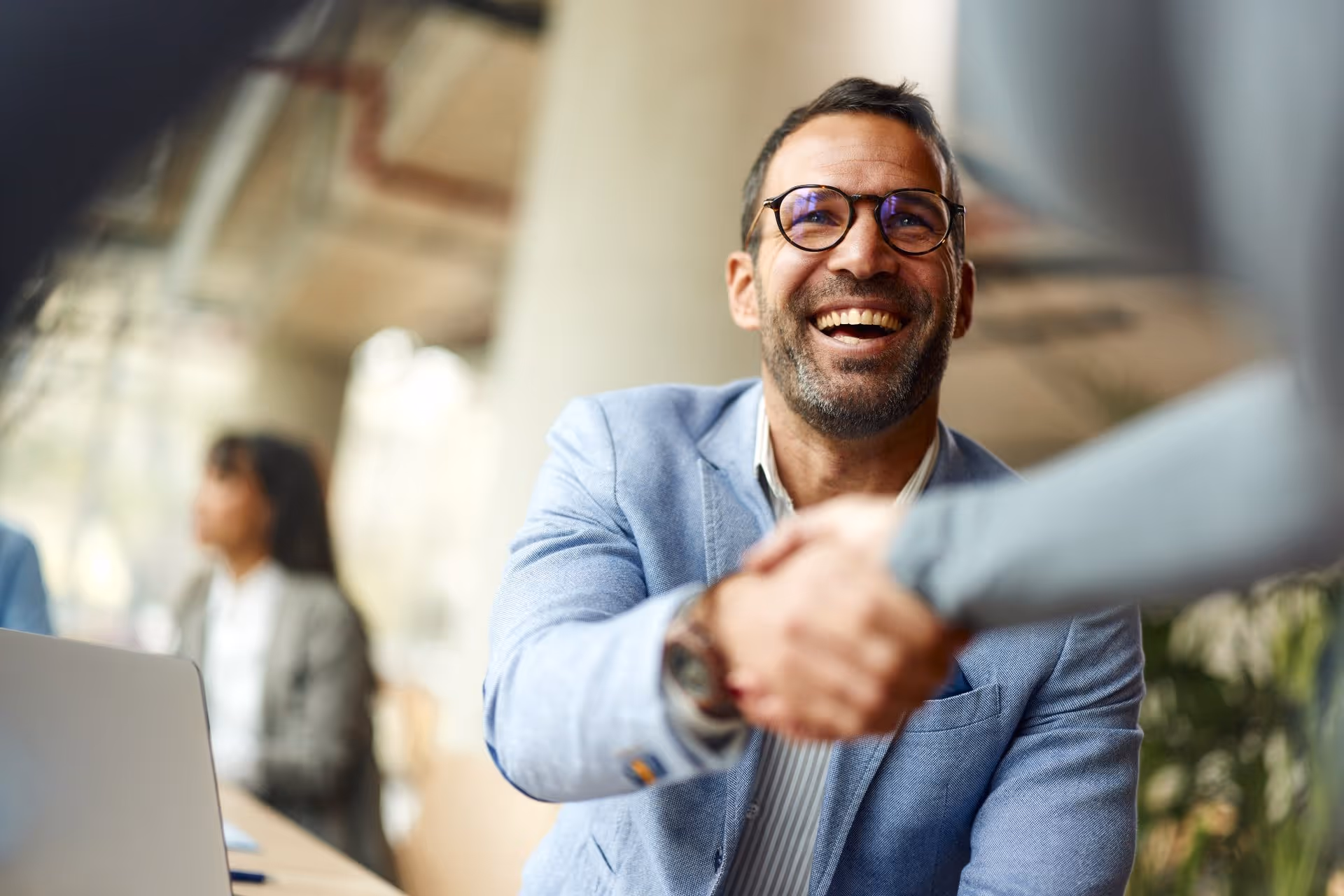 Smiling man in glasses and blue blazer shaking hands with someone in an office.