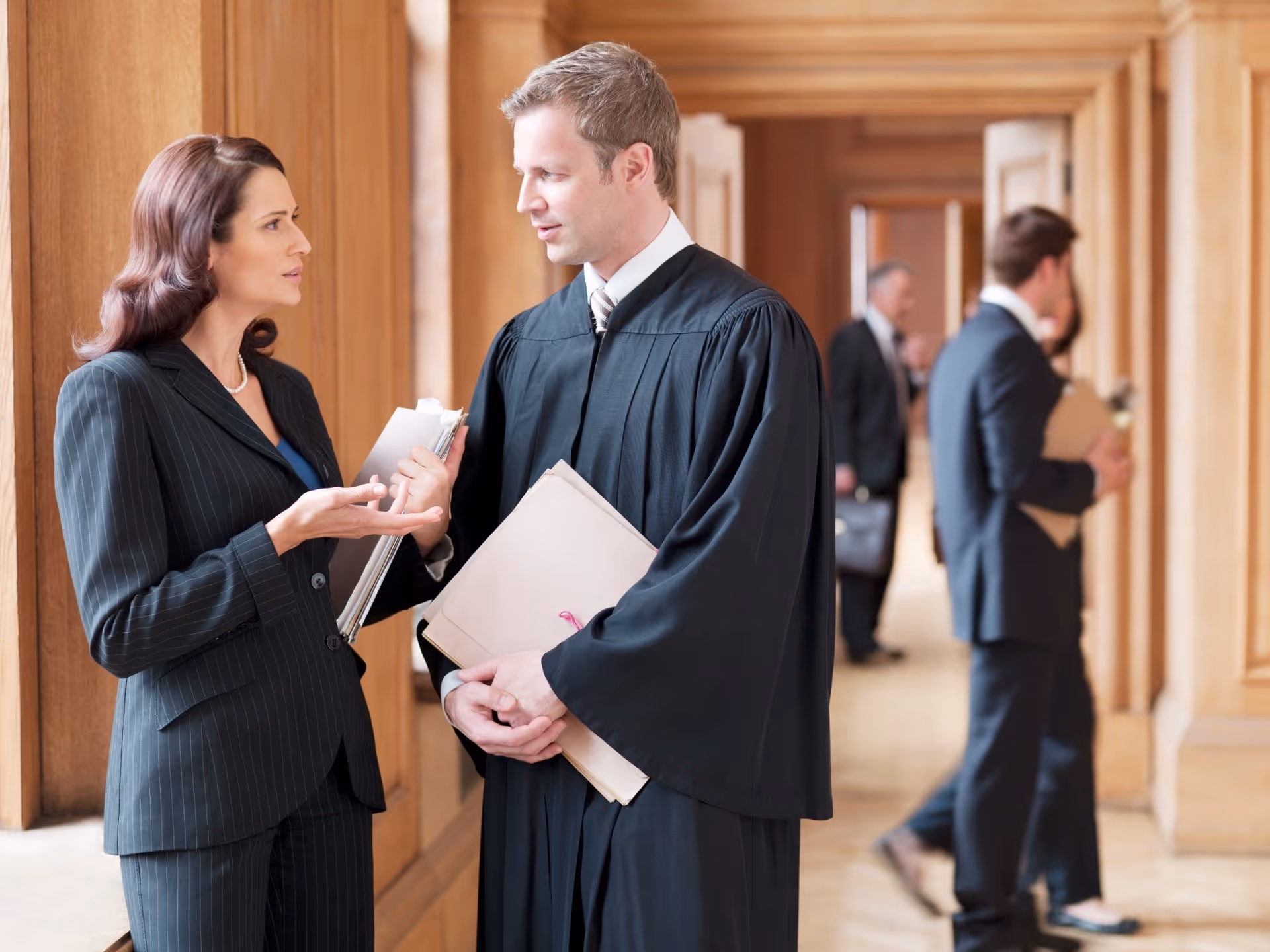 Man in judge robe holding files talking to woman in business suit in a courthouse hallway.