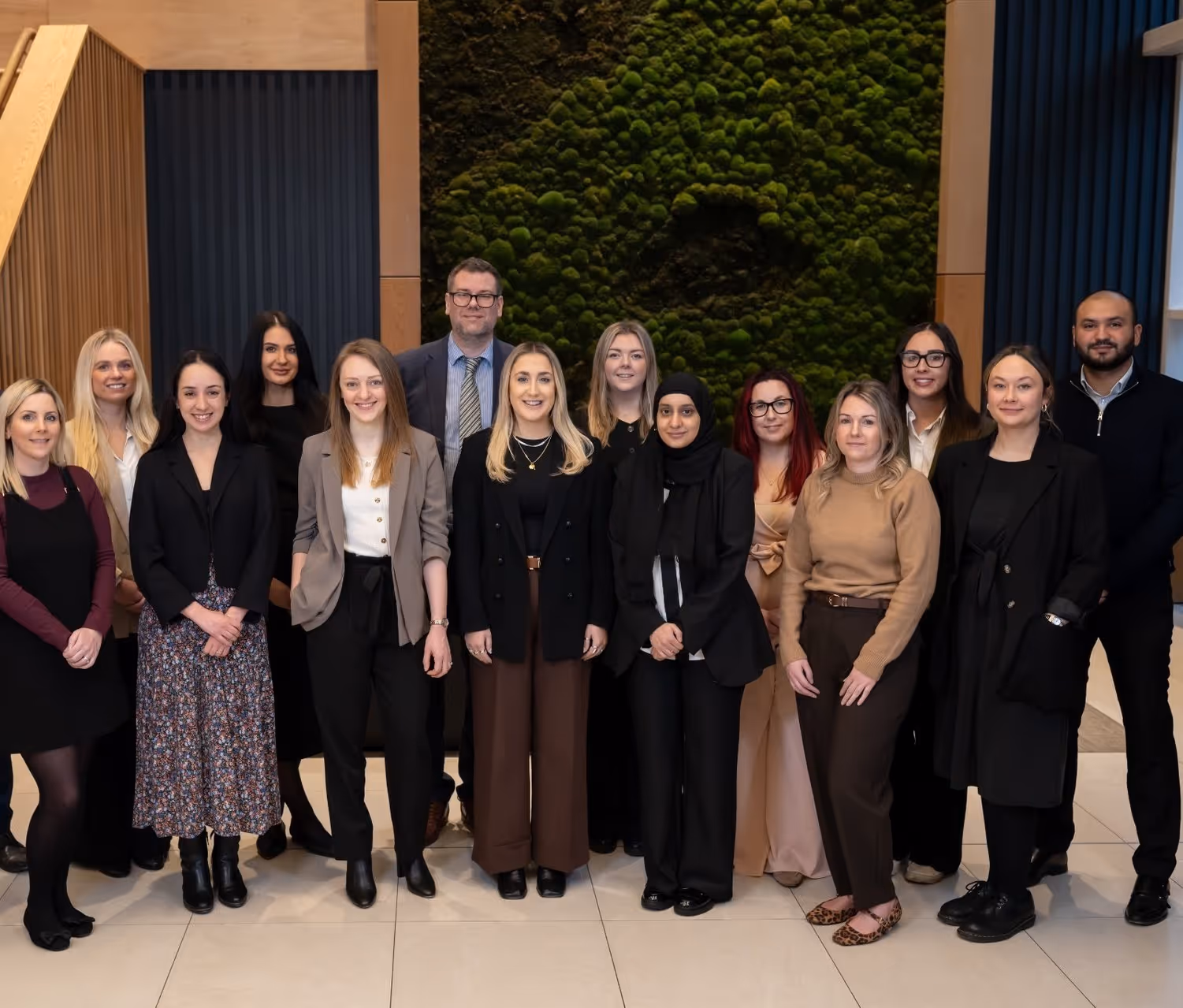 Group of 15 professionally dressed adults posing indoors in front of a moss wall installation.