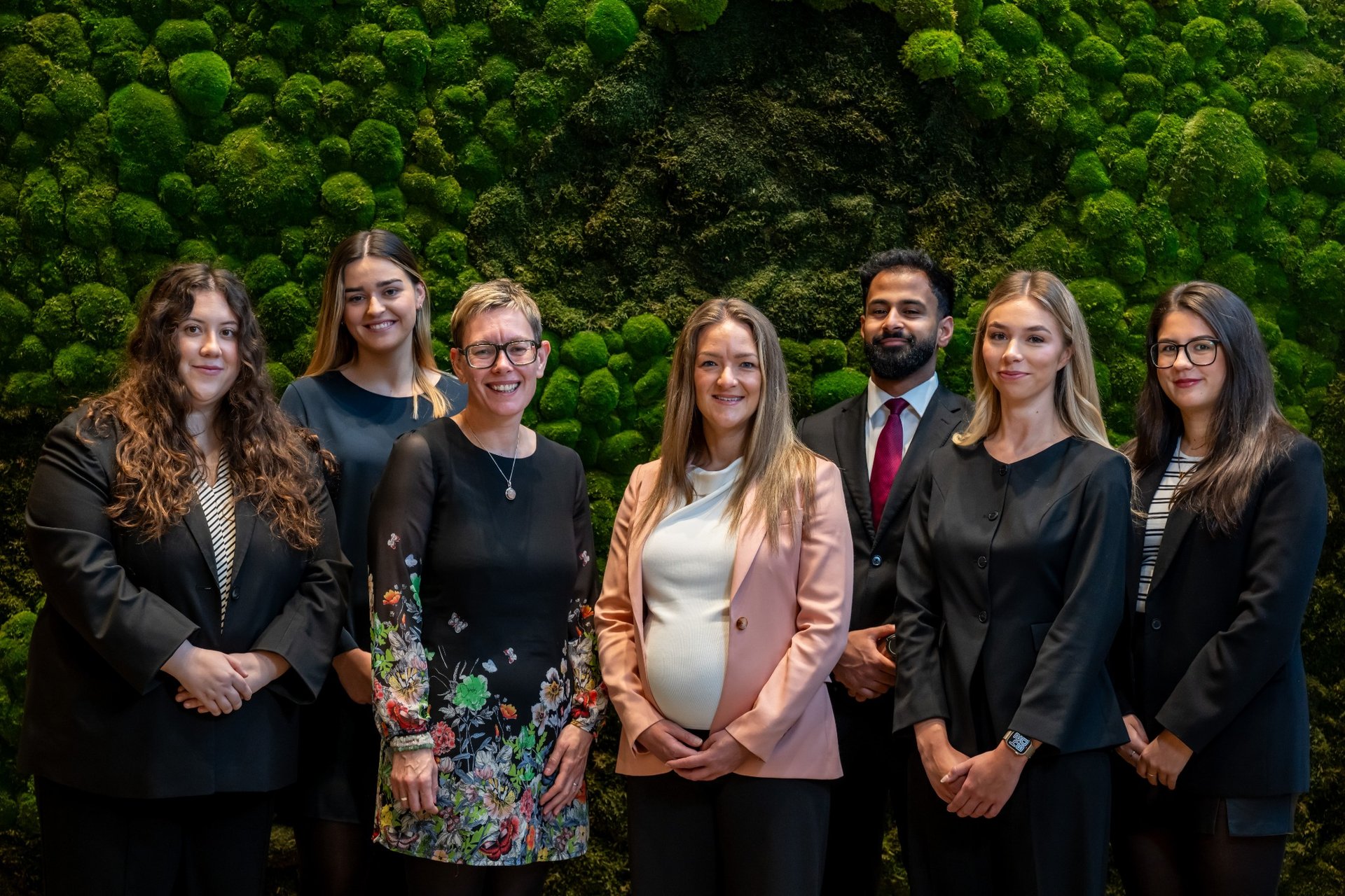 Group of six women and one man in business attire standing in front of a green moss wall.