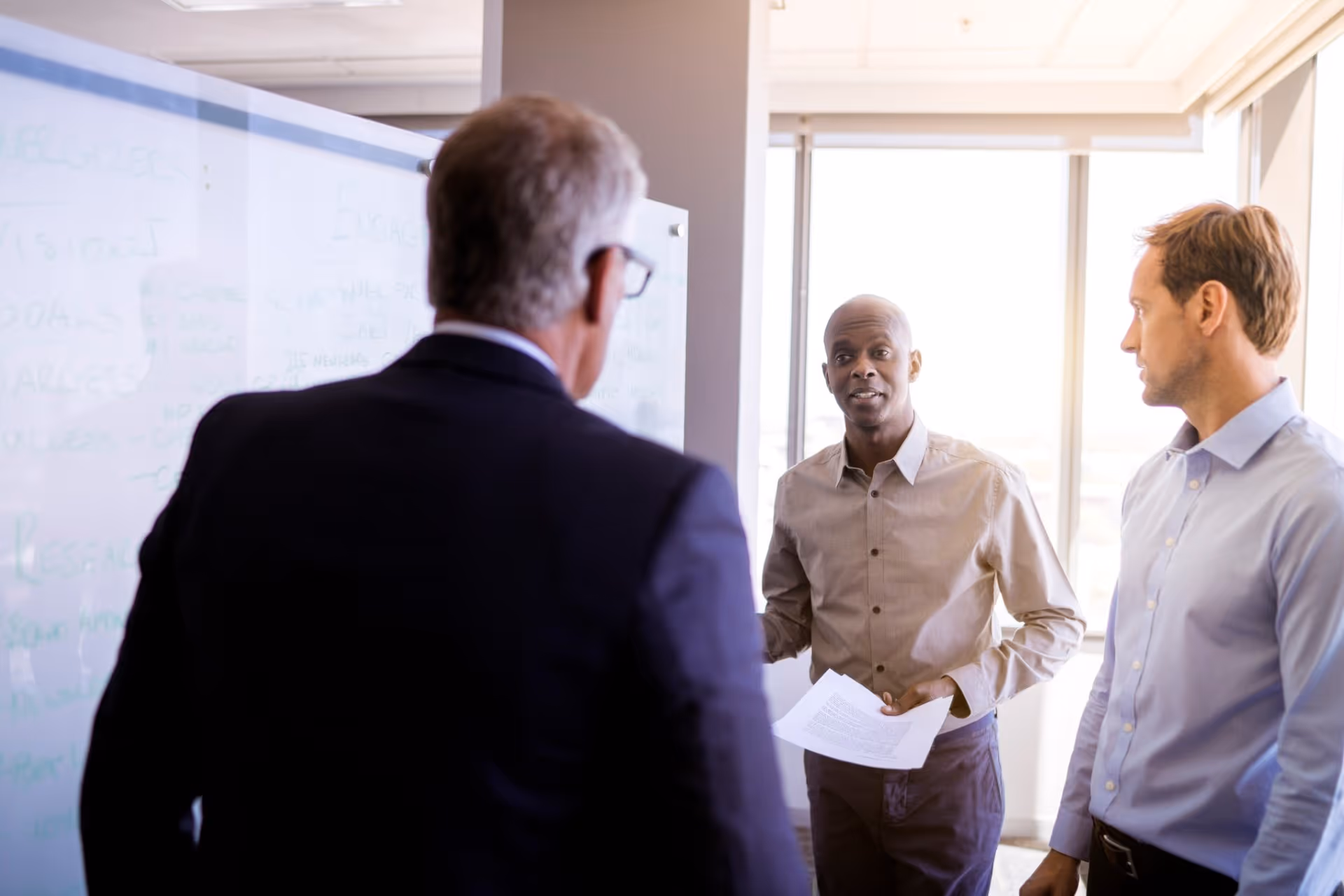 Three men discussing in a bright office beside a whiteboard with notes and papers in hand.
