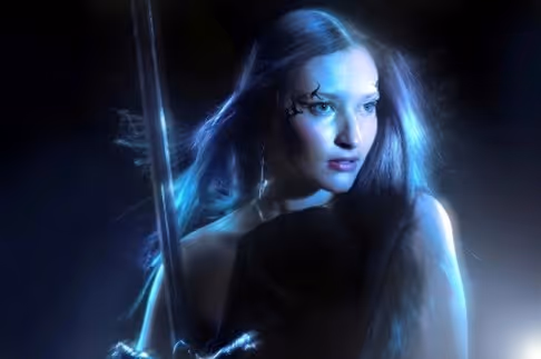 Woman with long hair and face paint holding a sword in glowing blue light.