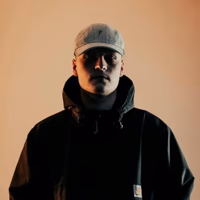 Portrait of a serious young man in a gray cap and dark jacket against a beige background.