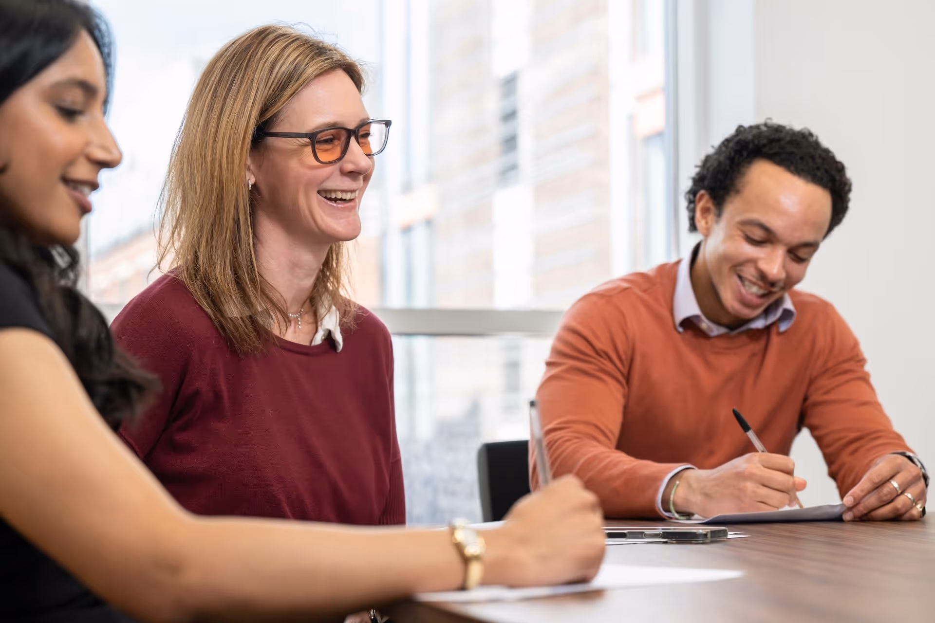 Three diverse people smiling and writing at a table in a bright office meeting.