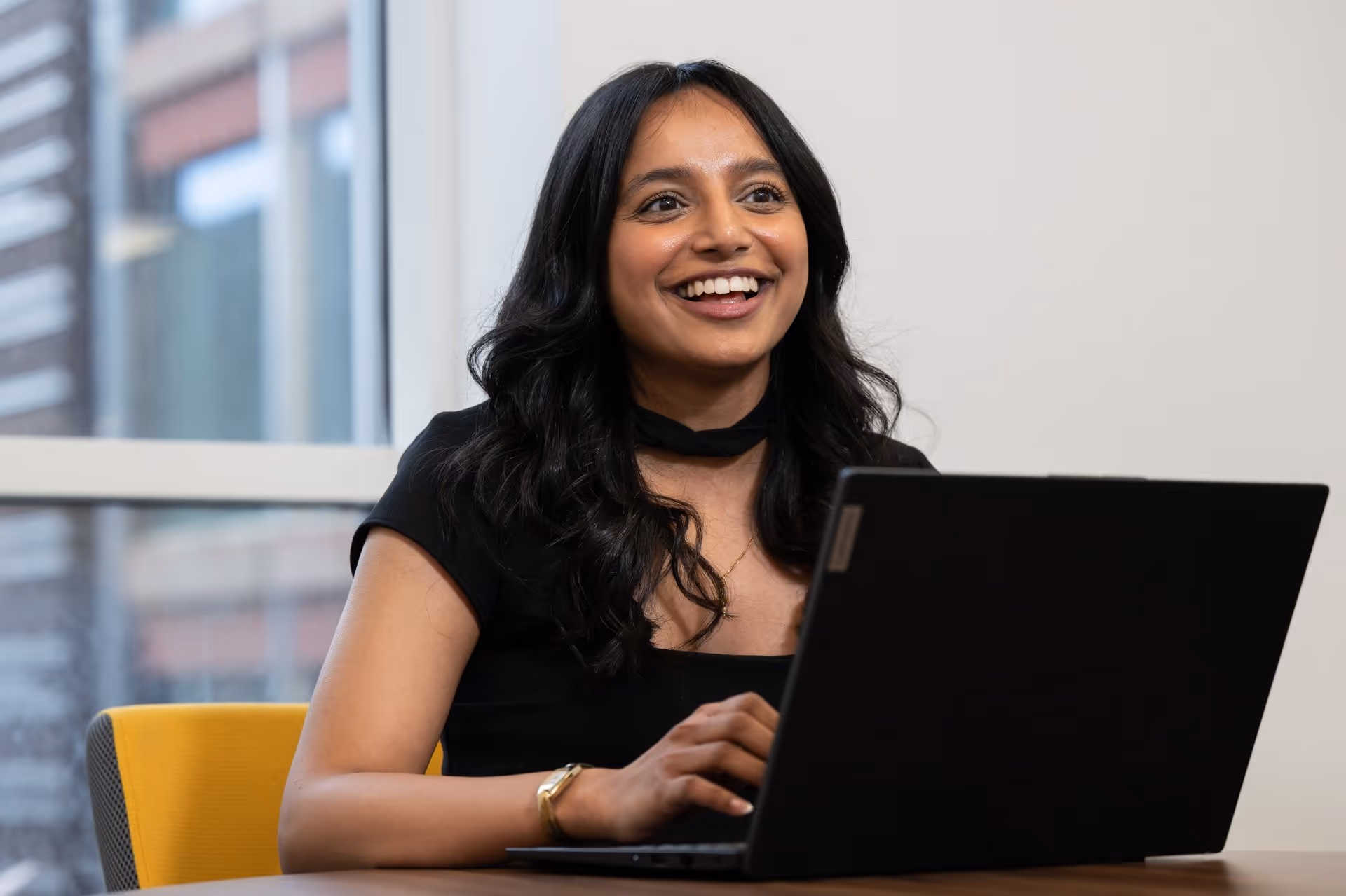 Smiling woman with black hair typing on a laptop at a desk near a window.
