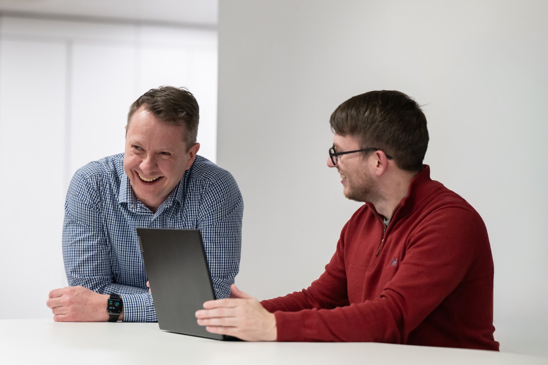 Two men sitting at a table, smiling and looking at each other, with a laptop in front of them.