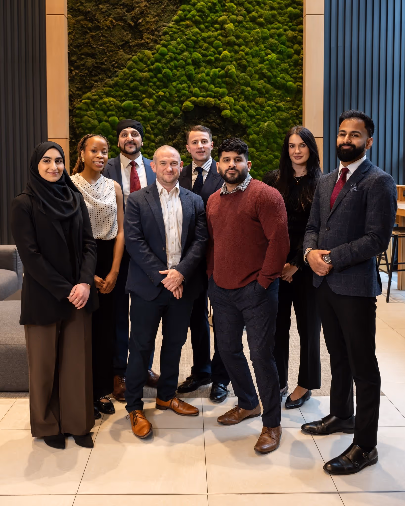 Group of eight diverse professionals standing indoors in business attire in front of a green moss wall.