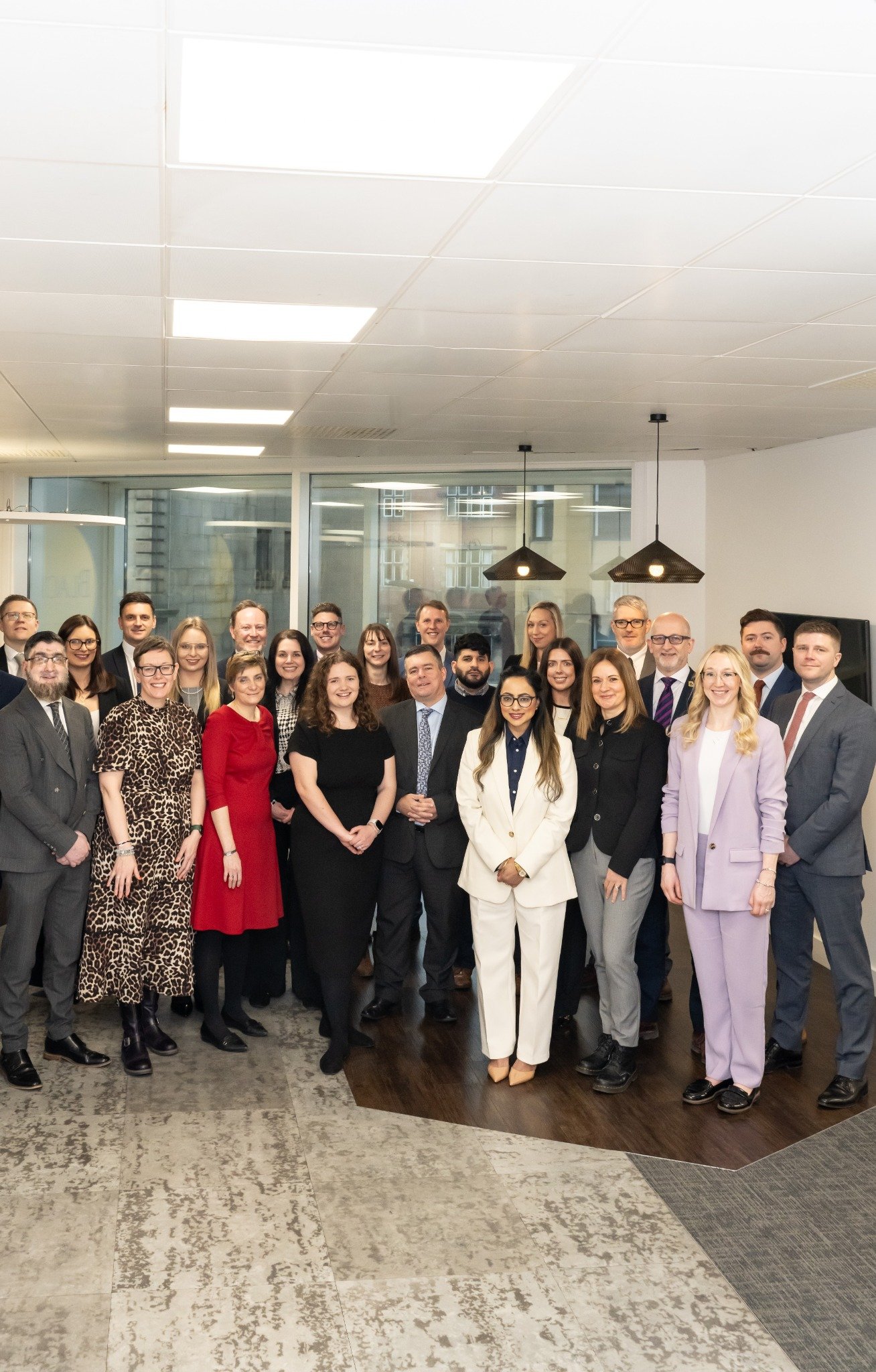 Group of 22 people in office setting wearing business attire, standing and smiling at camera.