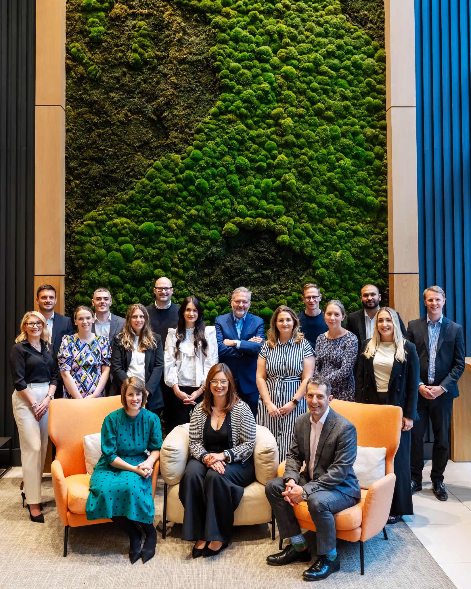 Group of 16 well-dressed people posing indoors in front of a green moss wall with two orange chairs.