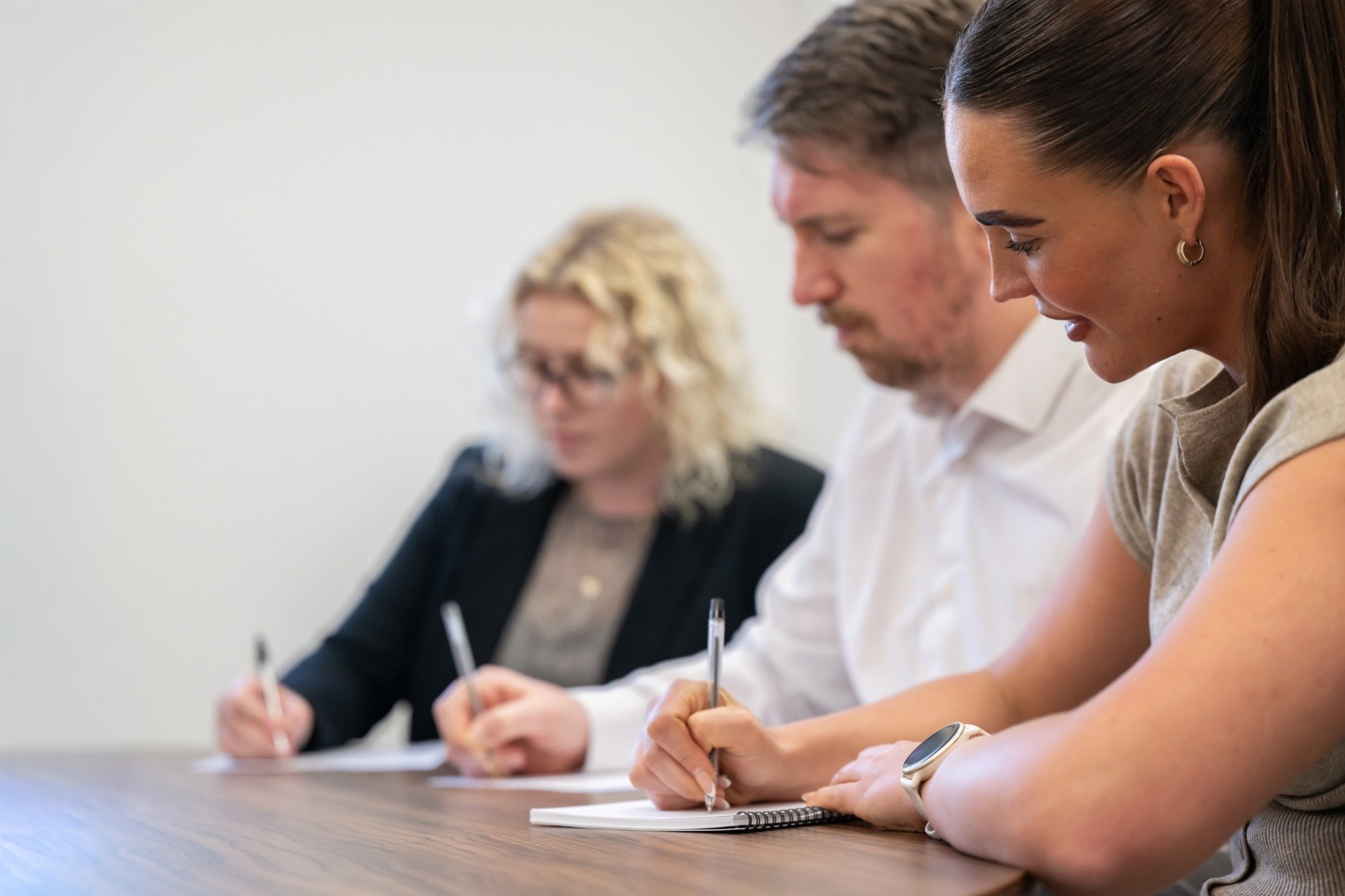 Three adults sitting at a wooden table writing notes with pens on notebooks or paper.