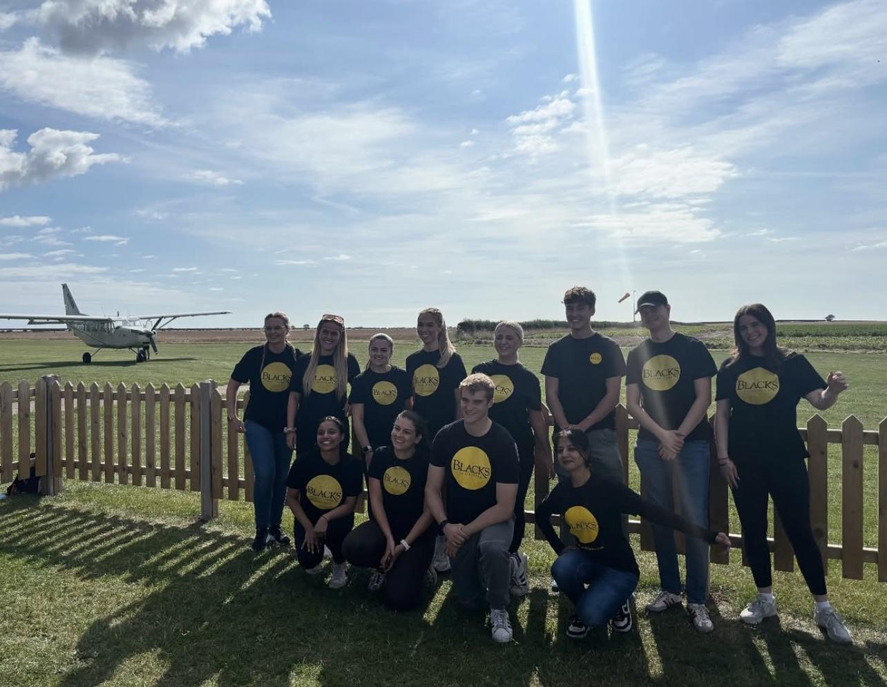 Group of people wearing black shirts with yellow logos posing outdoors near a small airplane and fence.