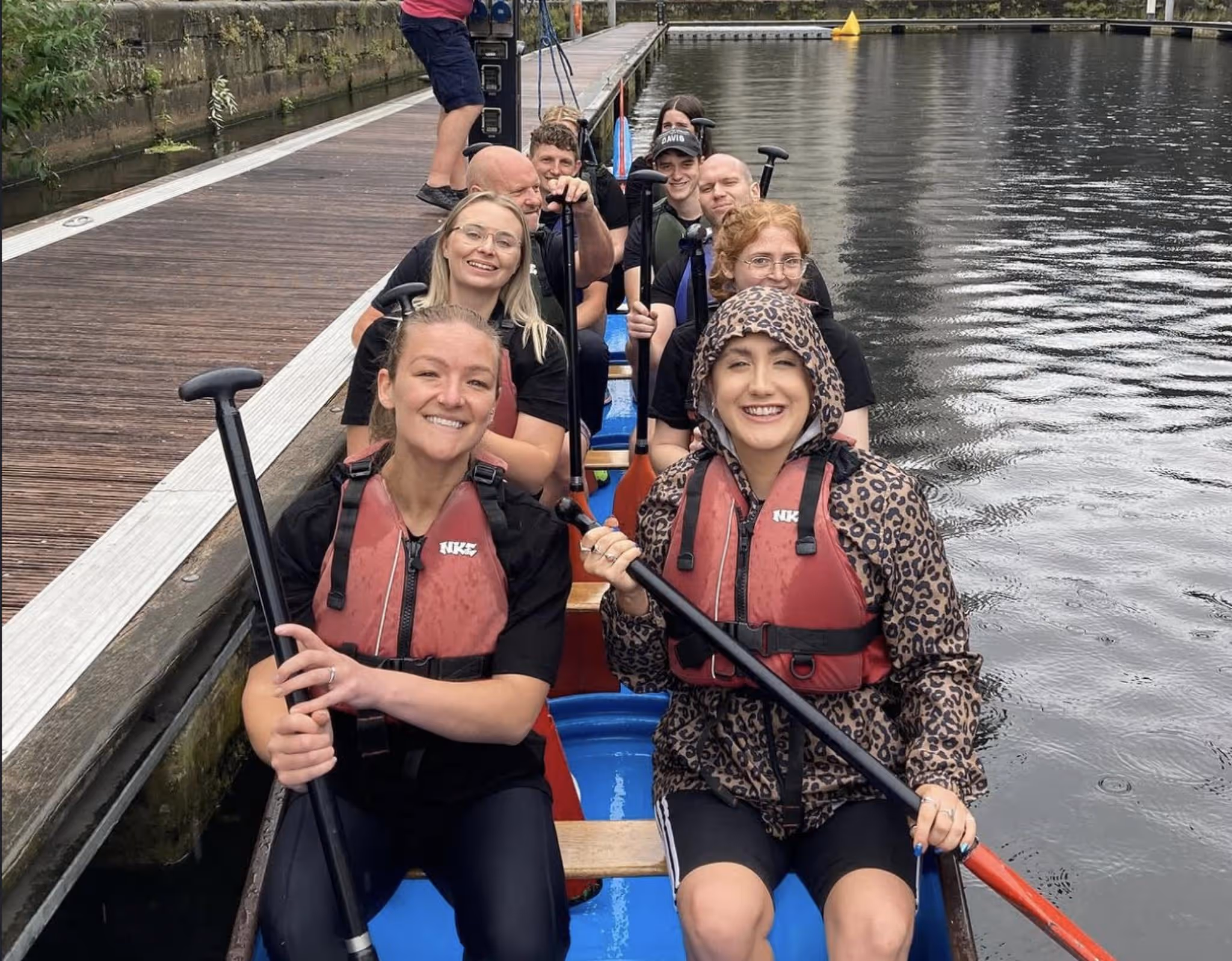 Smiling group of nine people in life jackets sitting in a blue canoe on water near a wooden dock.