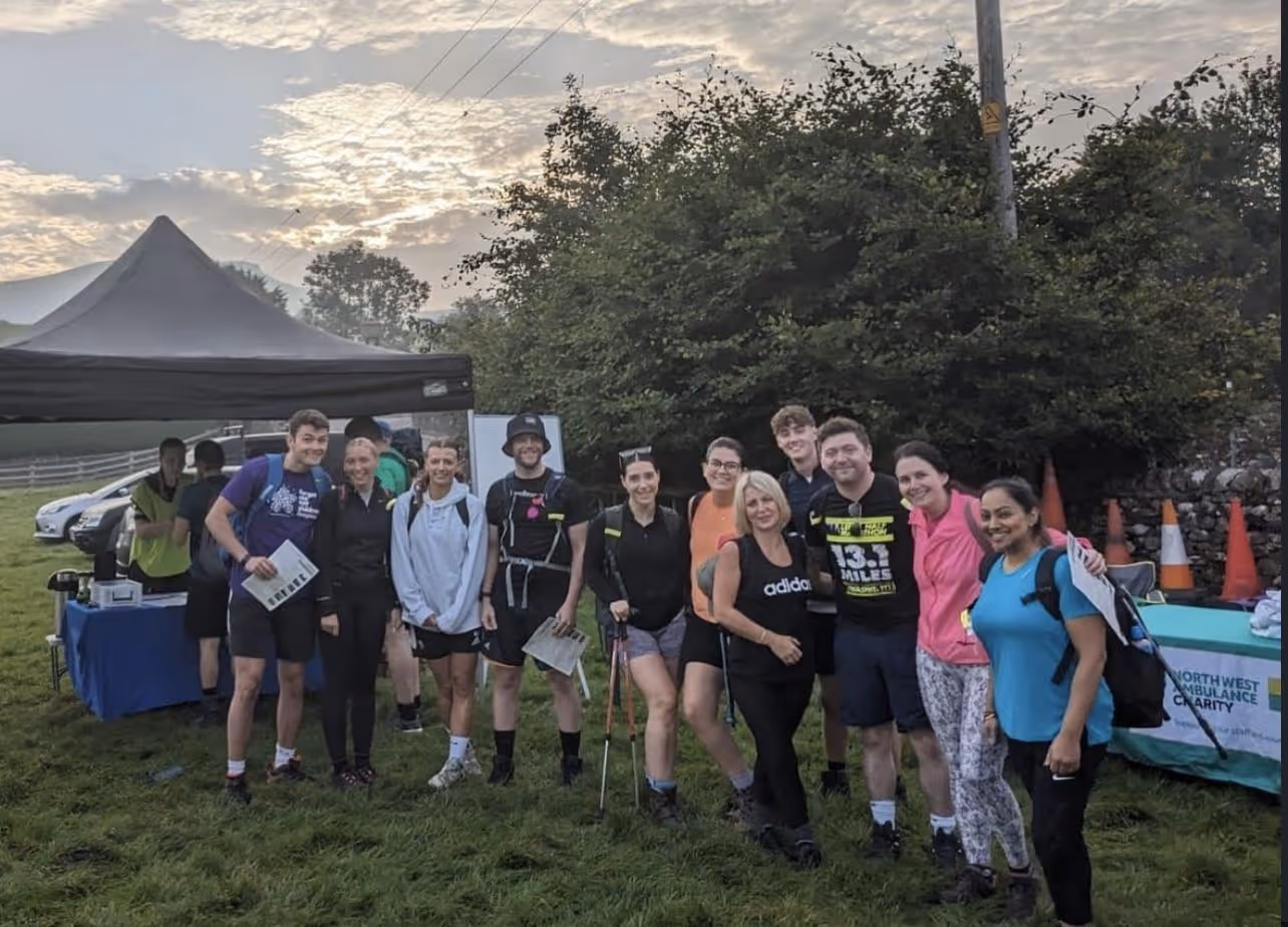 Group of smiling hikers standing on grass near tents and a charity table at sunrise or sunset.