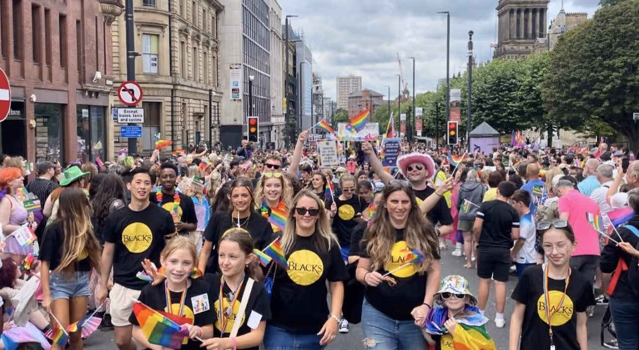 Crowd at a pride parade with people wearing black shirts holding rainbow flags on a city street.