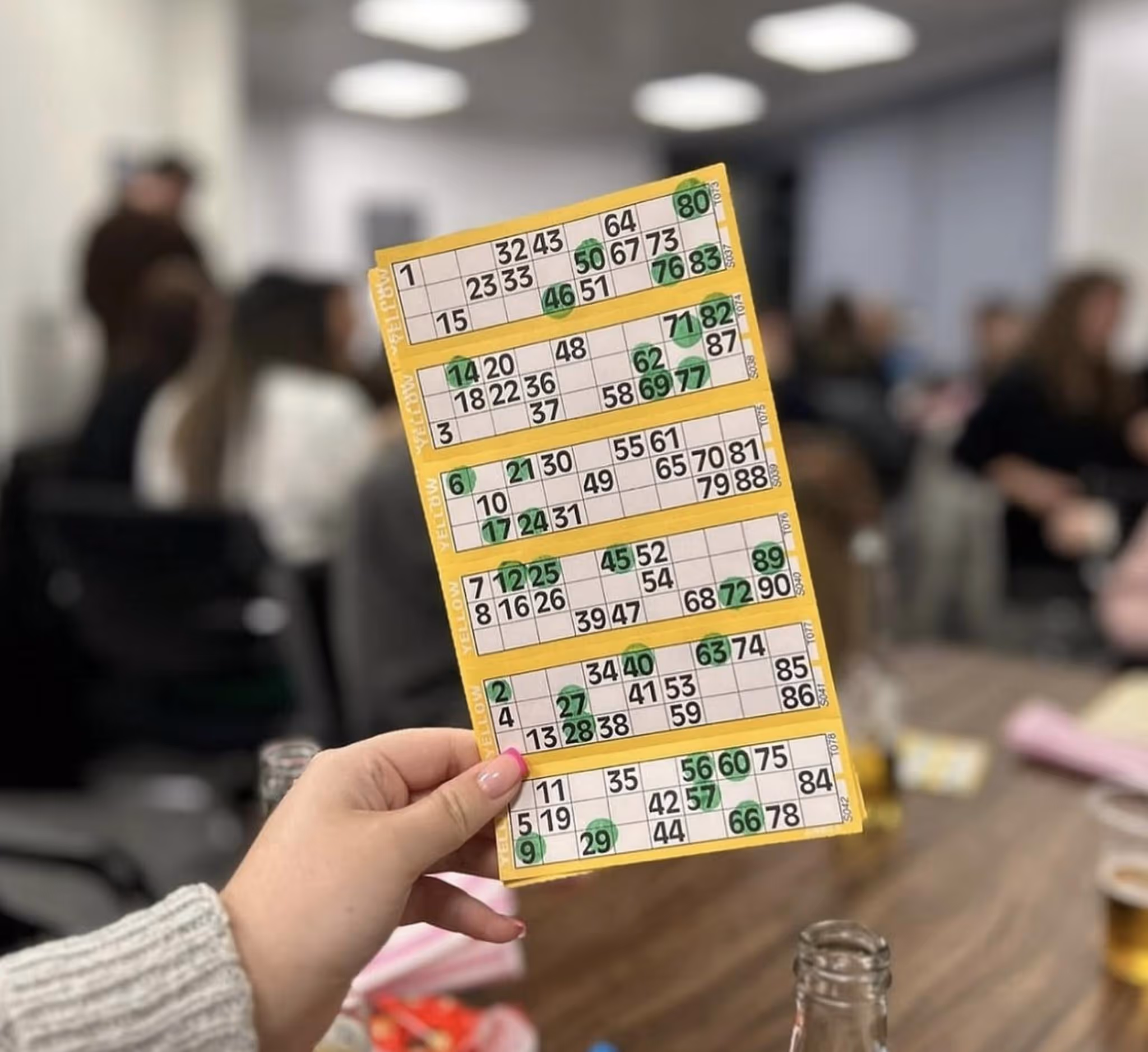 Hand holding a yellow bingo card with multiple green-marked numbers in an indoor setting with people.